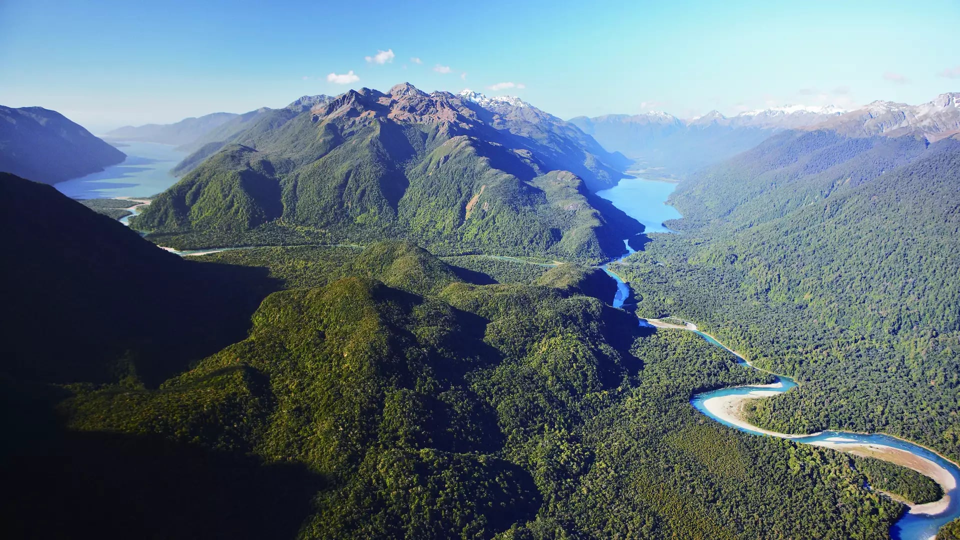 Aerial view of a valley with a winding river, forest, lakes, and mountains under a clear blue sky.