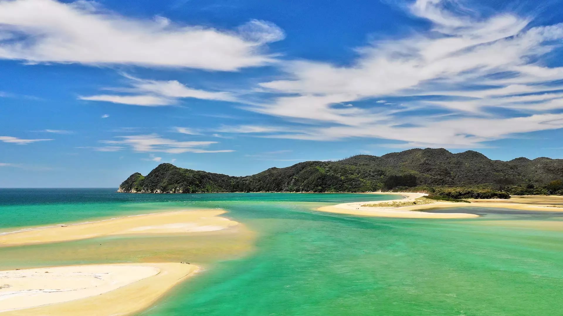 Sandy beach with turquoise water, green hills, and blue sky with clouds.
