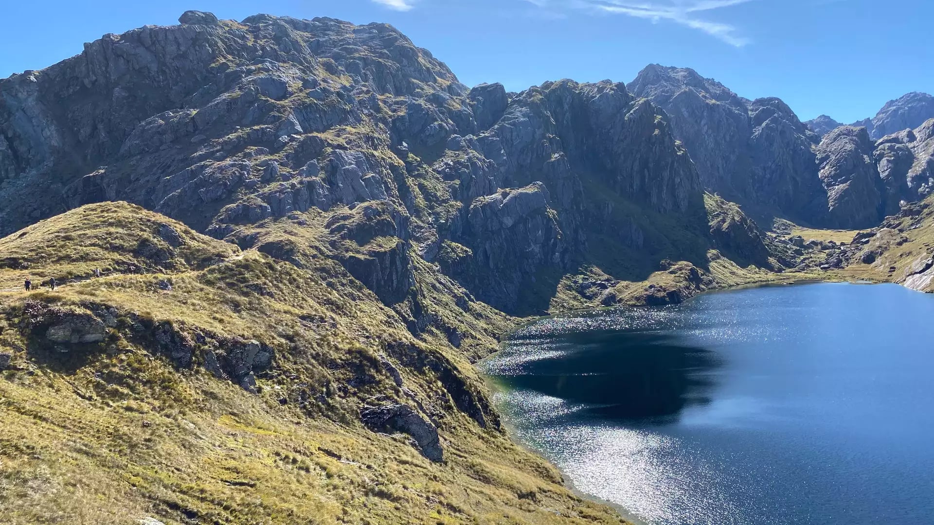 Mountain lake with rocky grassy slopes and hikers on a trail on a sunny day.
