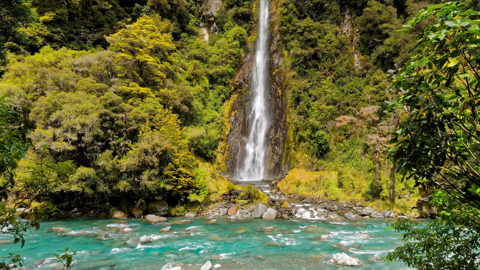 Tall waterfall cascading into bright blue river with green forest surrounding.