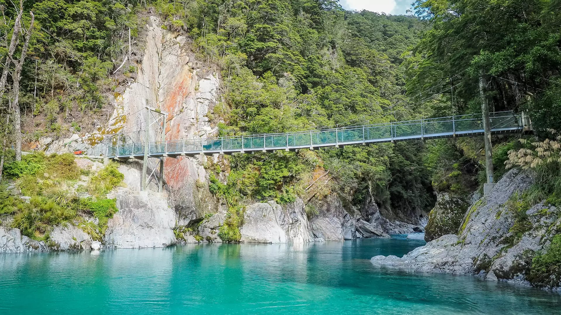 Suspension bridge over clear turquoise river with dense green forest and rocky cliffs