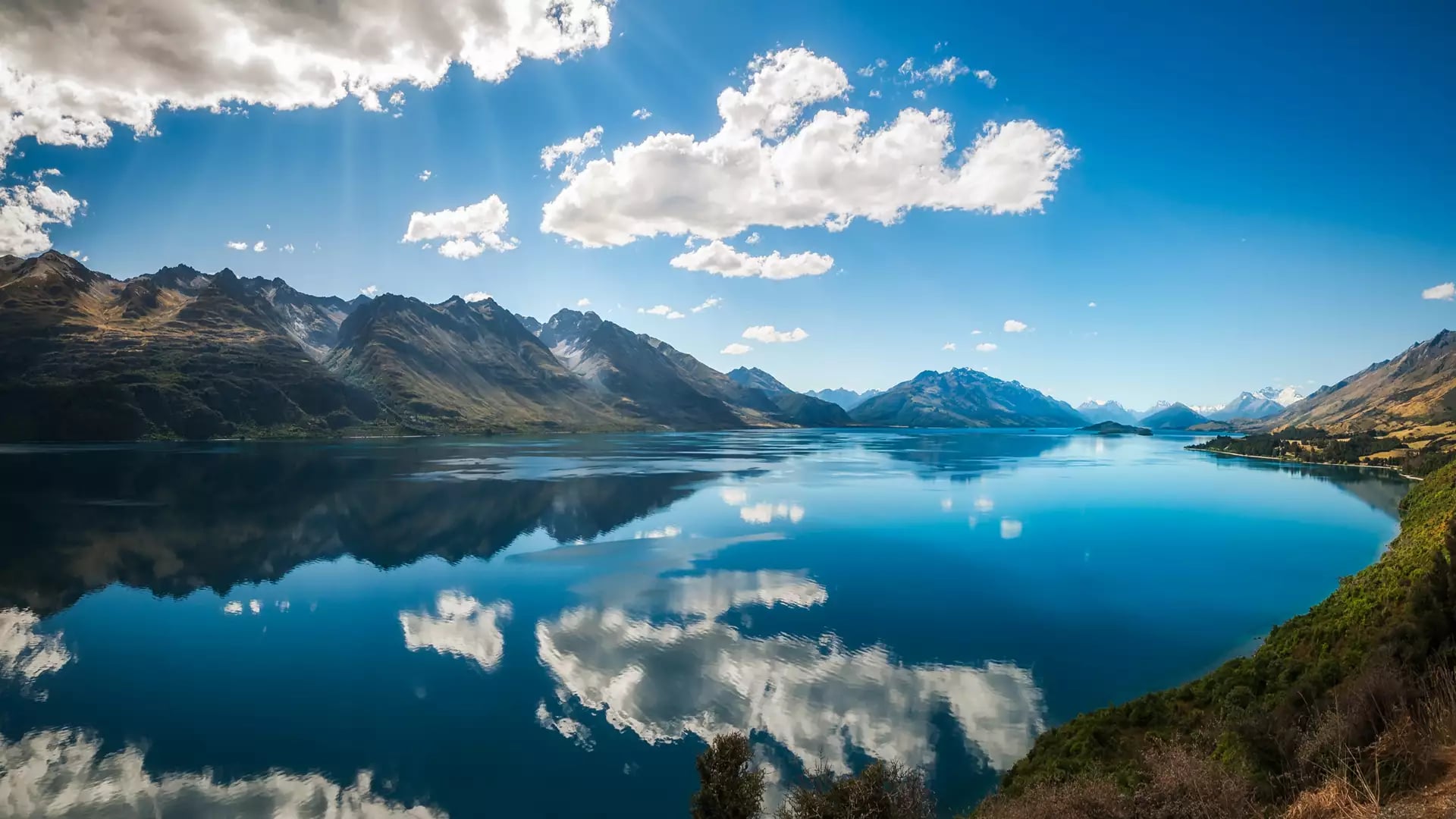 Clear lake reflecting rugged mountains and white clouds under a bright blue sky in Glenorchy.