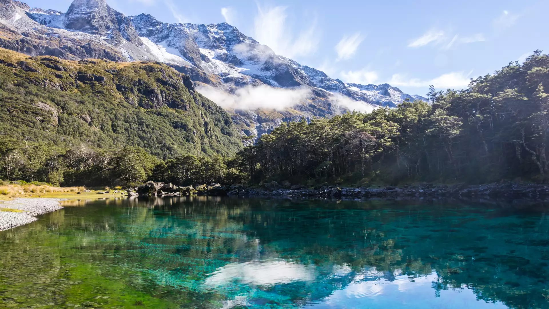 Clear lake with forest and snow-capped mountain reflections under blue sky