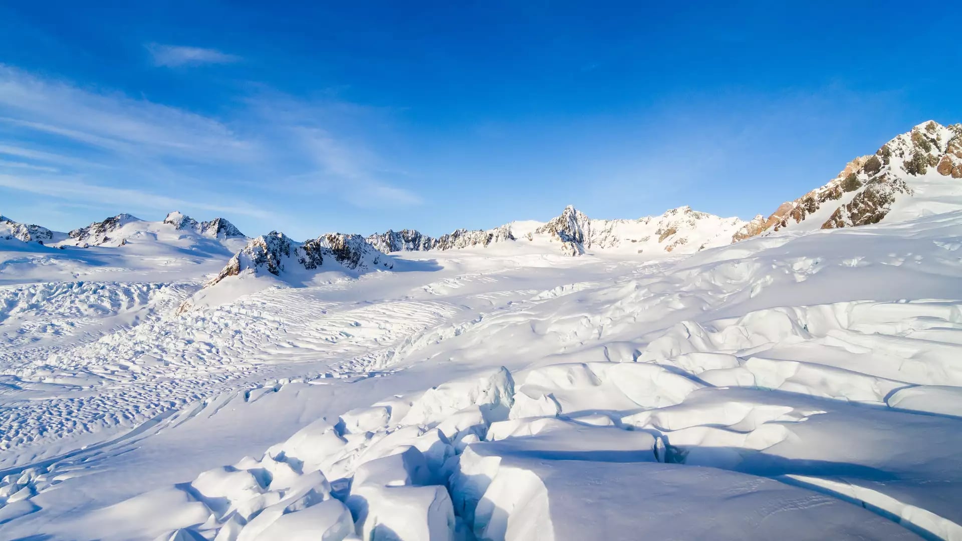 An expansive aerial view of the jagged ice falls and snow-covered névé of Fox Glacier under a bright blue sky.