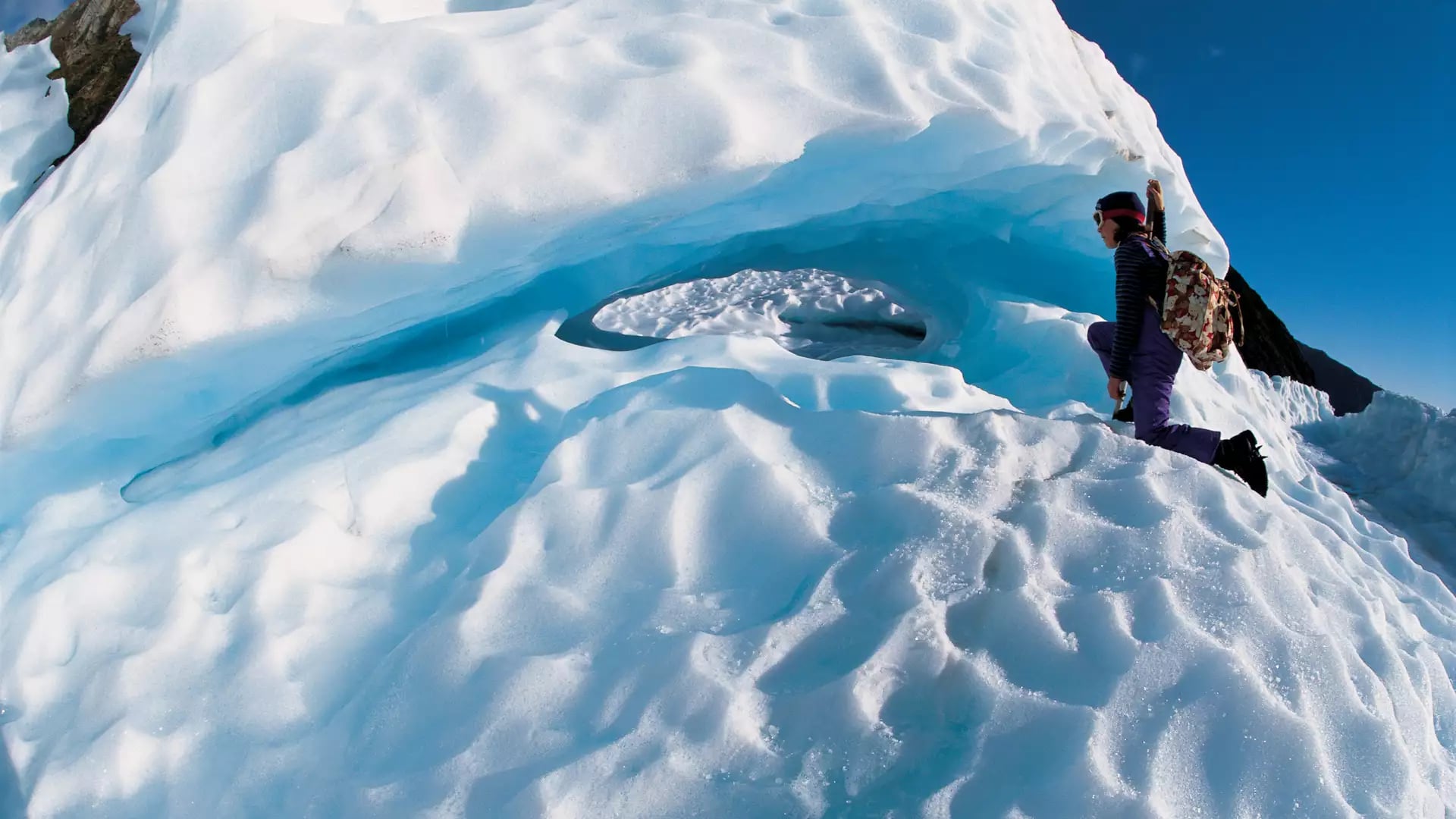 A hiker in purple gear and a backpack explores a glowing blue ice cave and dramatic ridges on the surface of Fox Glacier.