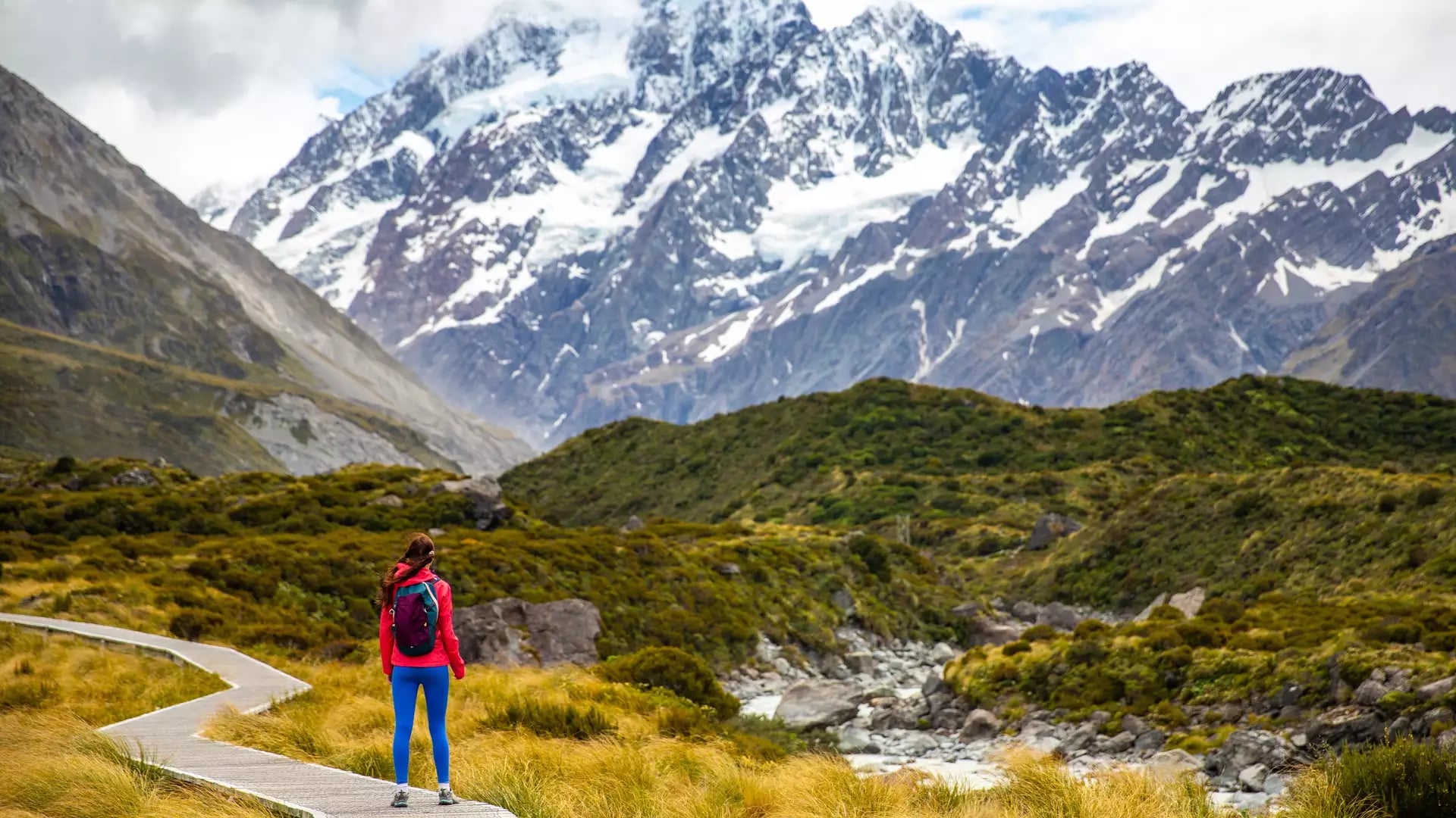 A person in bright colourful clothing standing on the walkway with Mt Cook in the background 