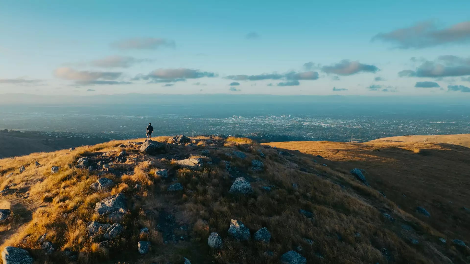 Person stands on rocky grassy hilltop looking over Christchurch city with clouds in blue sky.