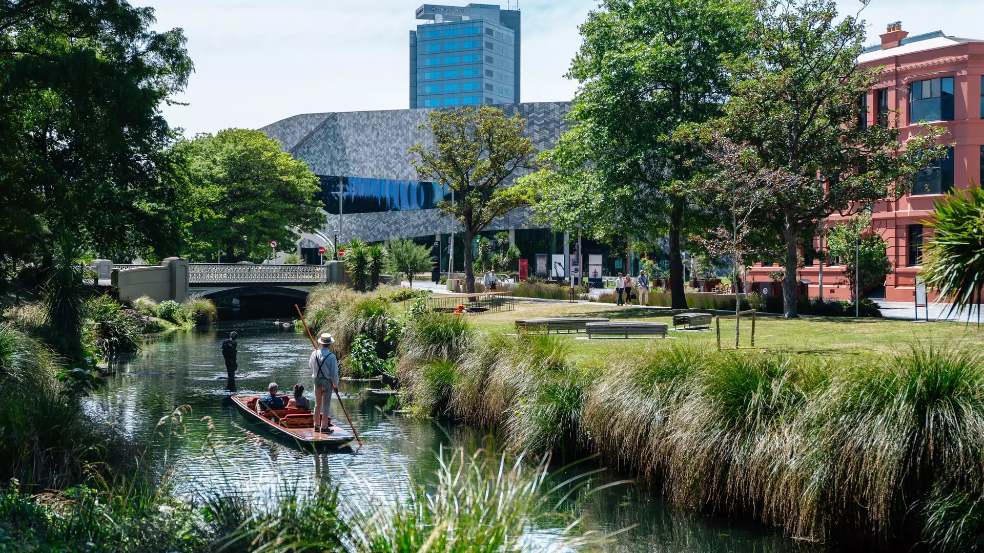People punting on a calm river with green trees, grass, and urban buildings in Christchurch.