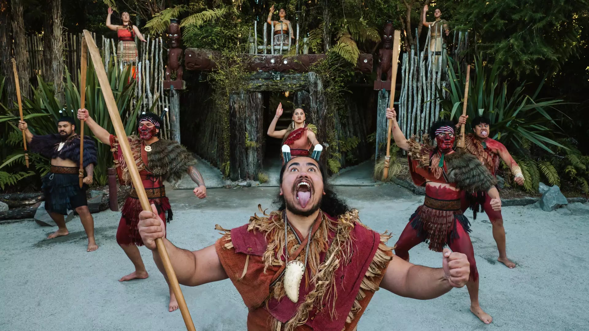 Maori kapa haka group performing traditional dance with spears and face paint in forest setting.