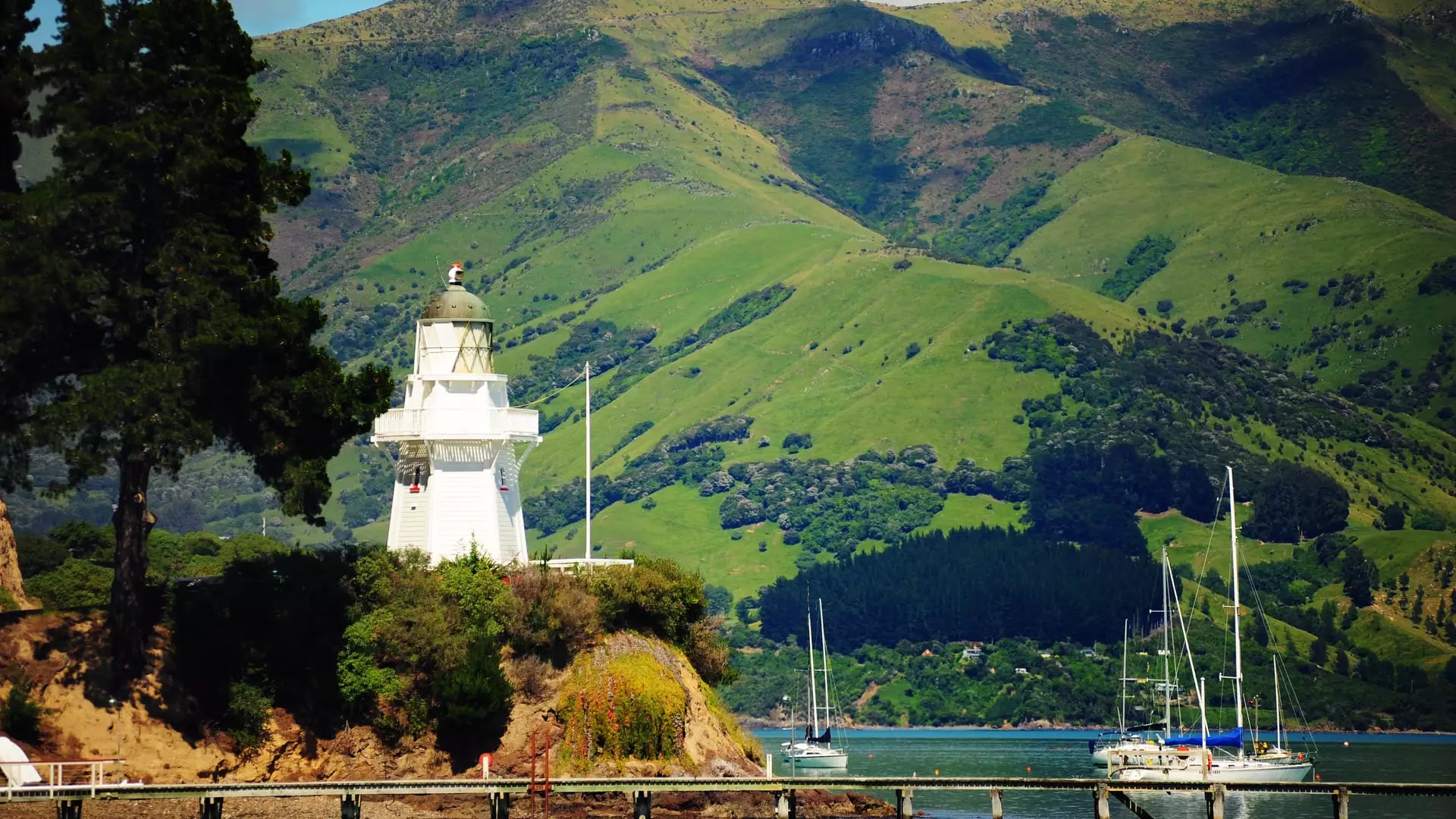 White lighthouse on a coastal hill with boats on the water and green hills in the background.