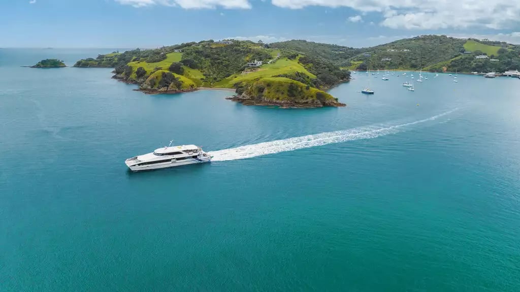 Ferry boat sailing near green hills of Waiheke Island with other boats anchored in calm blue water.