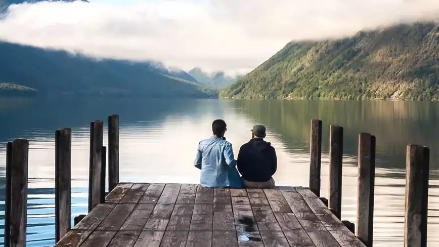 Two people sitting on a dock at Nelson Lake Rotoiti with misty hills.