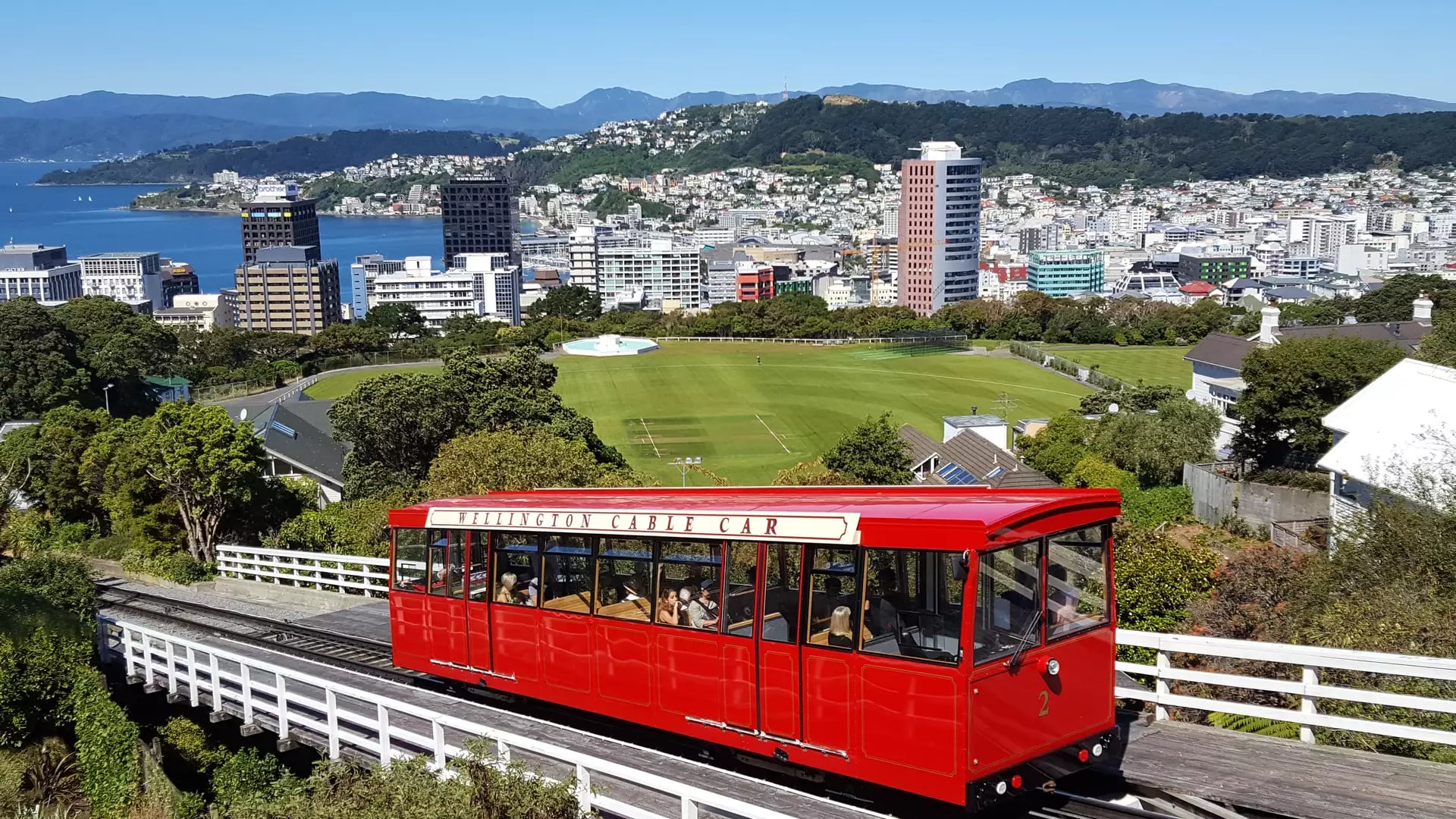 The bright red Wellington Cable Car travels down its tracks overlooking Wellington city, the harbor, and the surrounding hills under a clear blue sky.