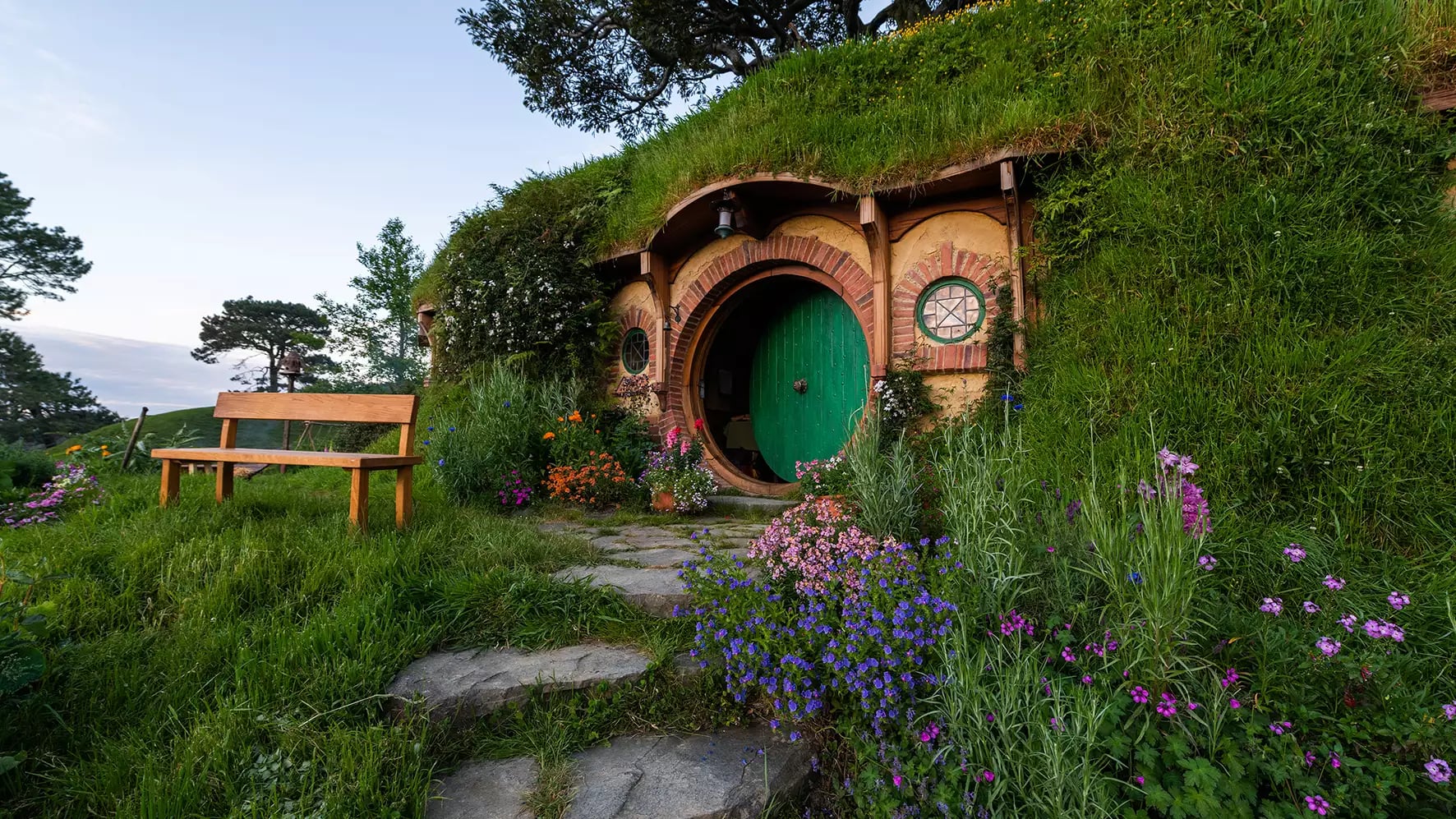 Hobbit hole with a green round door, surrounded by flowers and a wooden bench outside.
