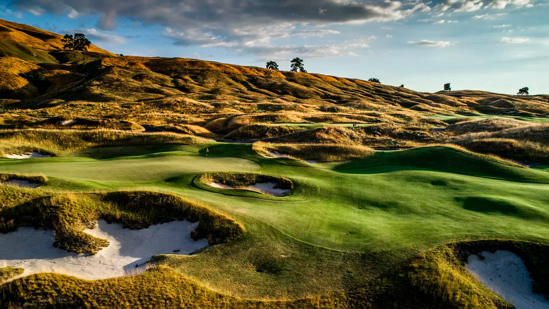 Golf course fairway with green, sand bunkers, and hills under cloudy sunset sky.