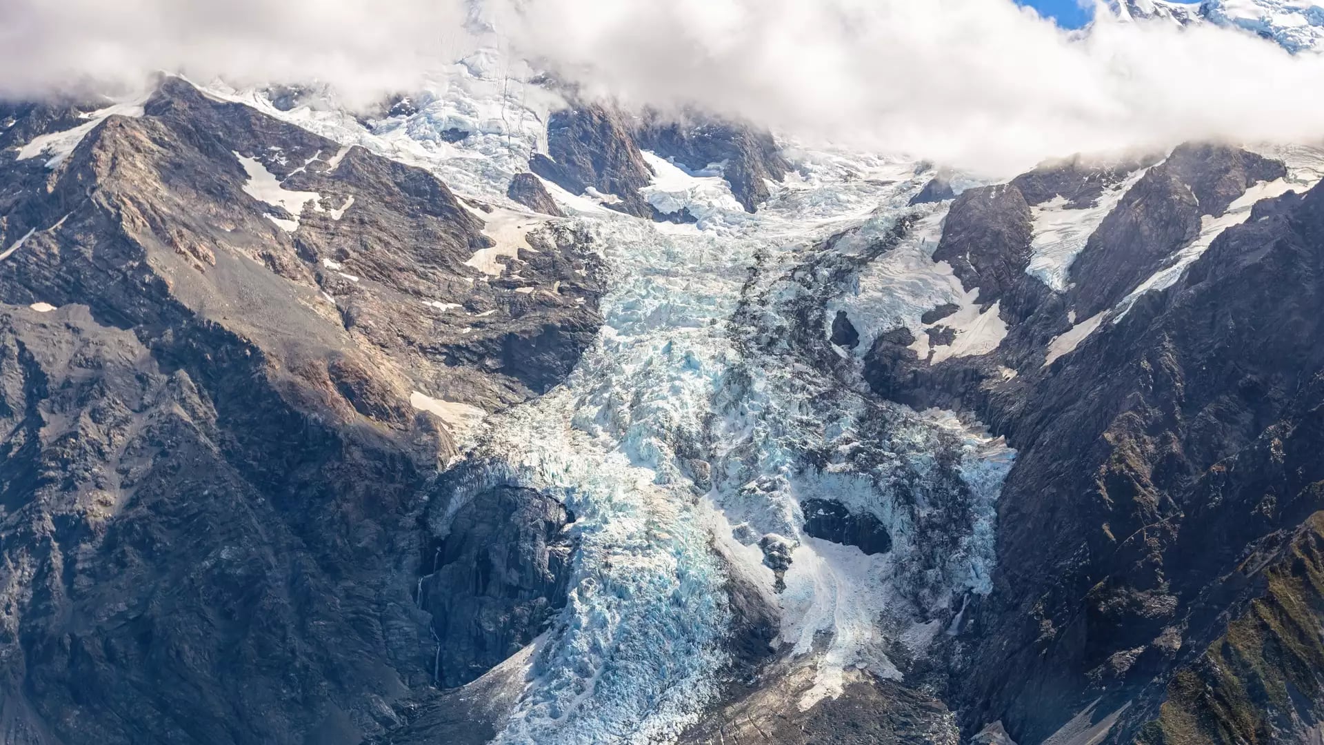 Snow-capped Fox Glacier and rocky mountains under blue sky with white clouds.