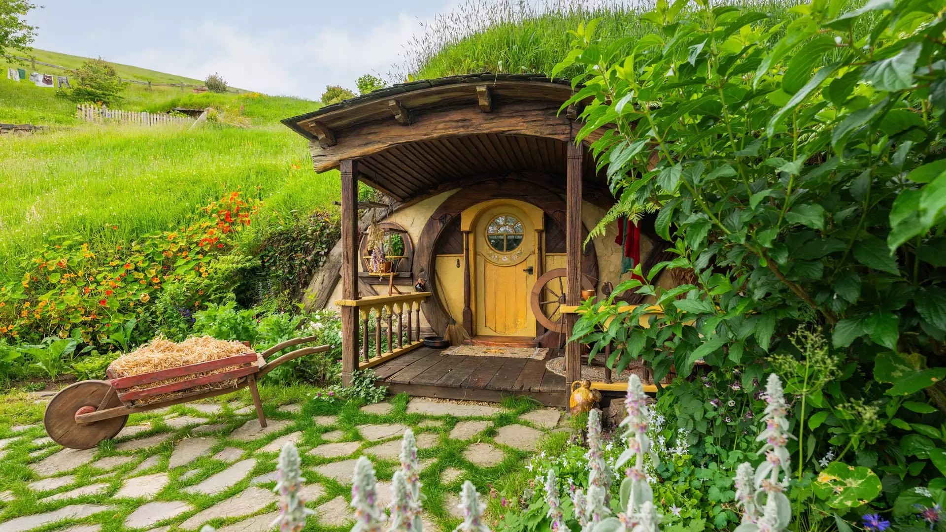 Hobbit house with round yellow door and wooden porch surrounded by greenery and flowers with a wheelbarrow in front.