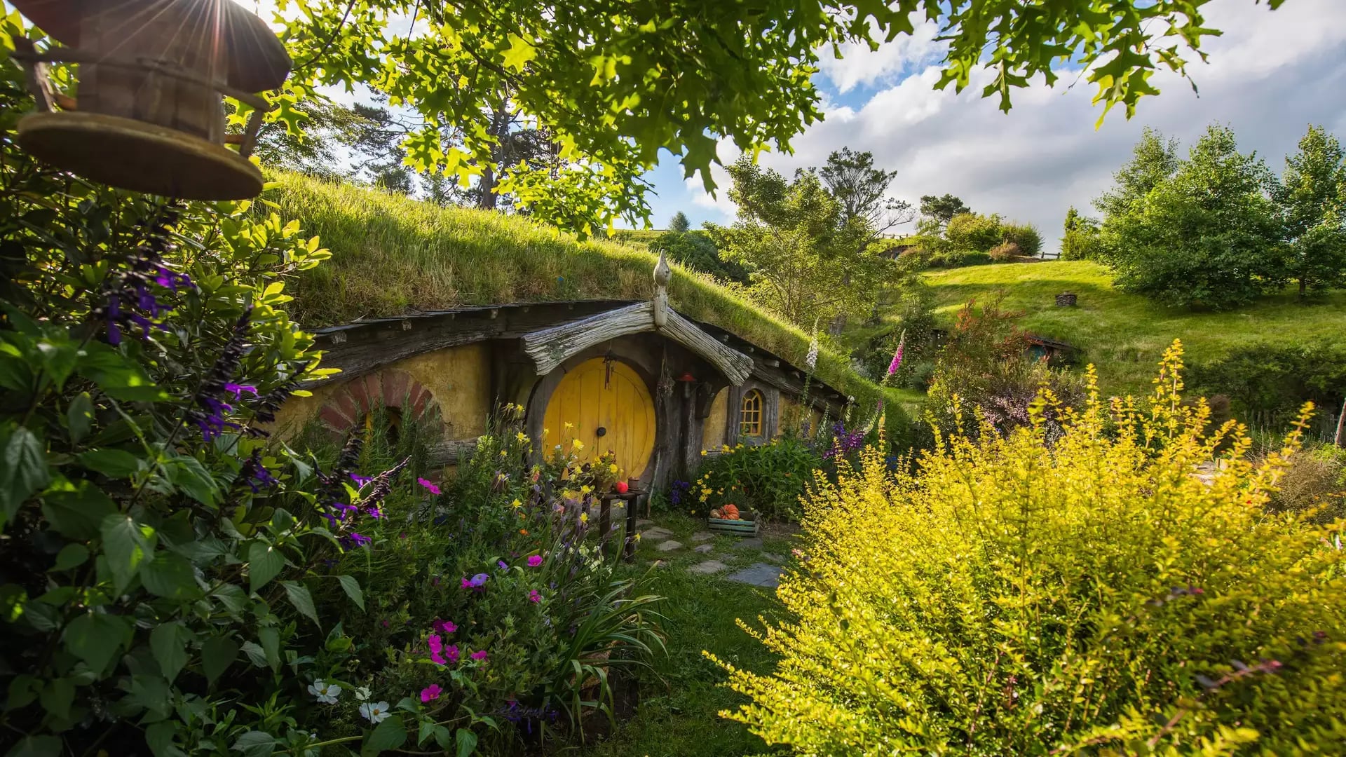 Hobbit hole with yellow door surrounded by vibrant garden and greenery under sunny sky.