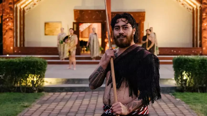 Maori warrior in traditional attire with wooden spear in front of a meeting house.