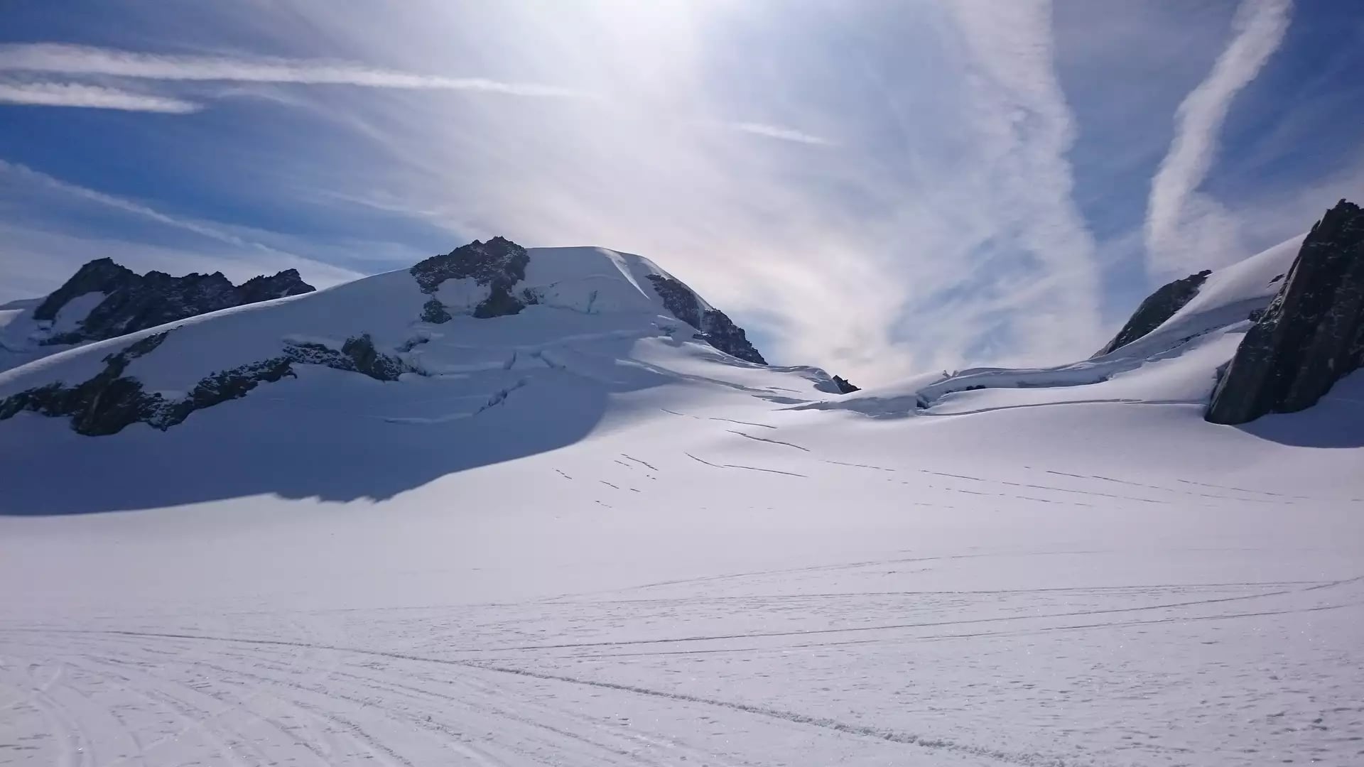 A vast, pristine snowfield on the upper reaches of Franz Josef Glacier, with deep crevasses visible on the mountain slopes under a bright sun and wispy clouds.