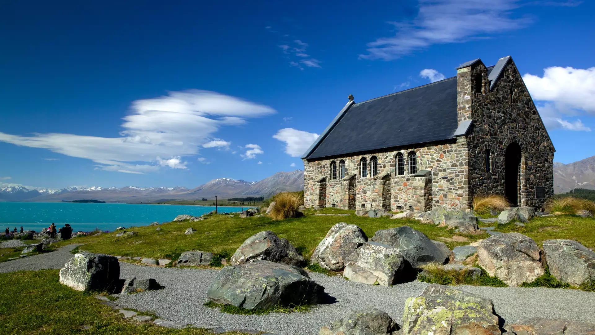 Stone church near turquoise lake with mountains and blue sky in the background.