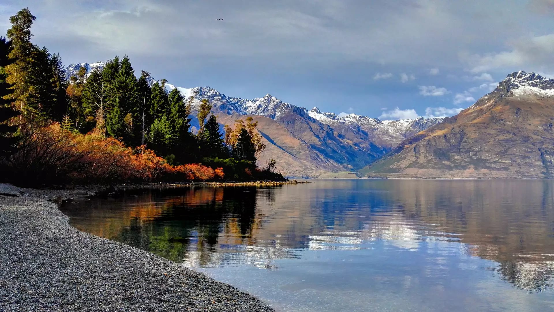 Lake with clear water reflecting snow-capped mountains and trees on a partly cloudy day.