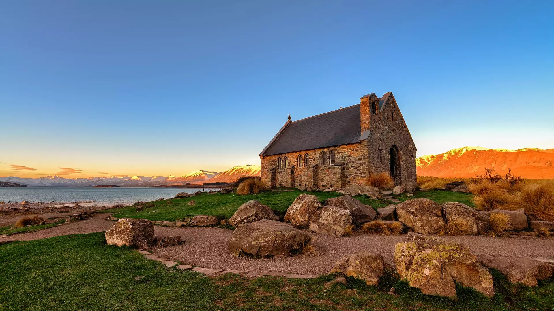 Stone church at sunset near a lake with mountains in the background.