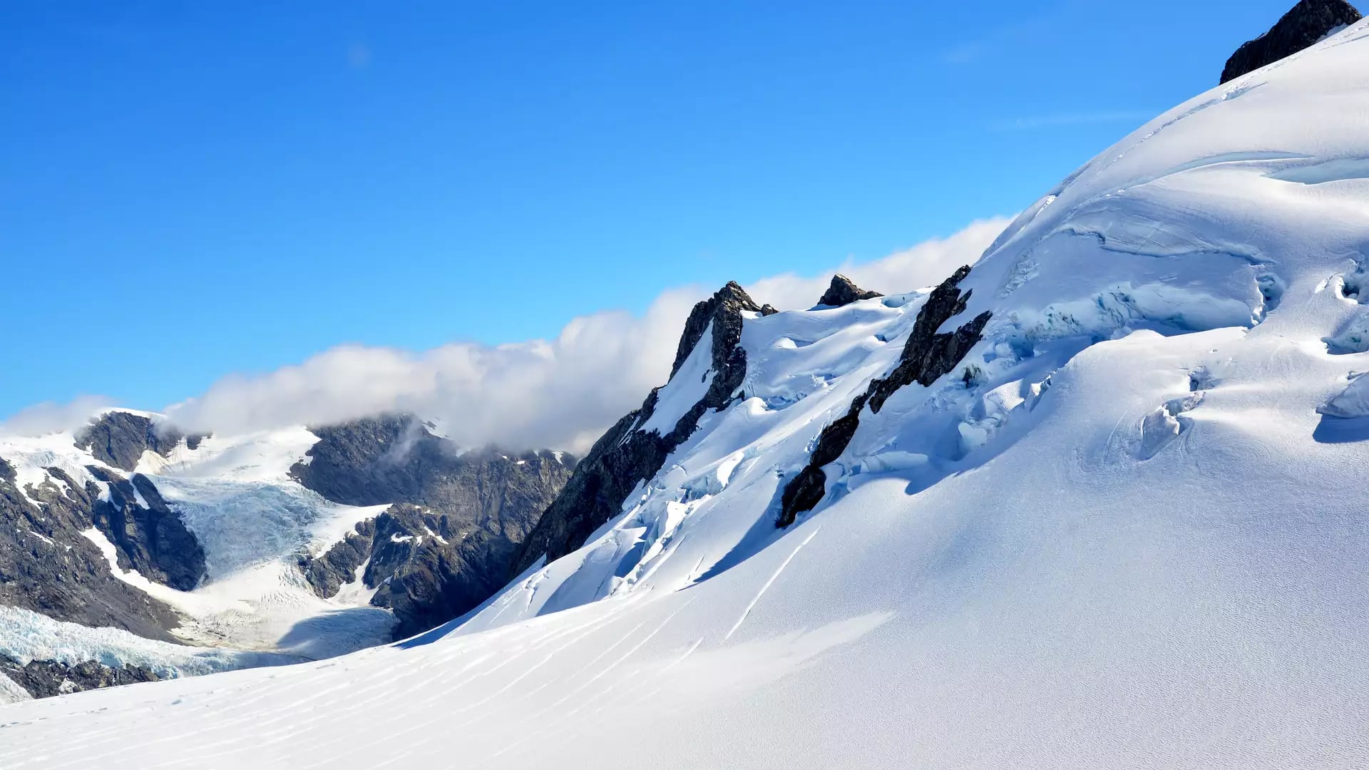 A bright, sunlit view of a steep, snow-covered mountain ridge with blue ice crevasses, set against a vibrant blue sky in the Southern Alps.