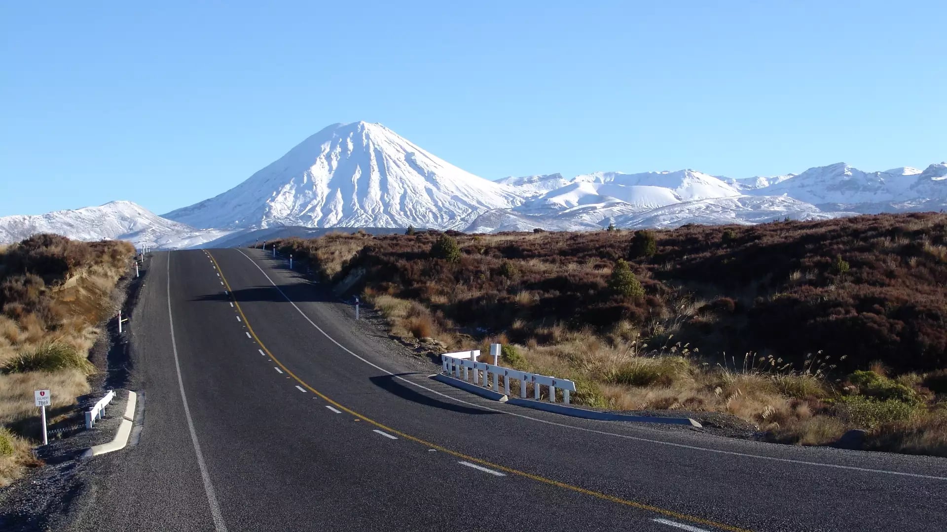 Paved road heading towards snow-covered Mount Ngauruhoe volcano under a clear blue sky.