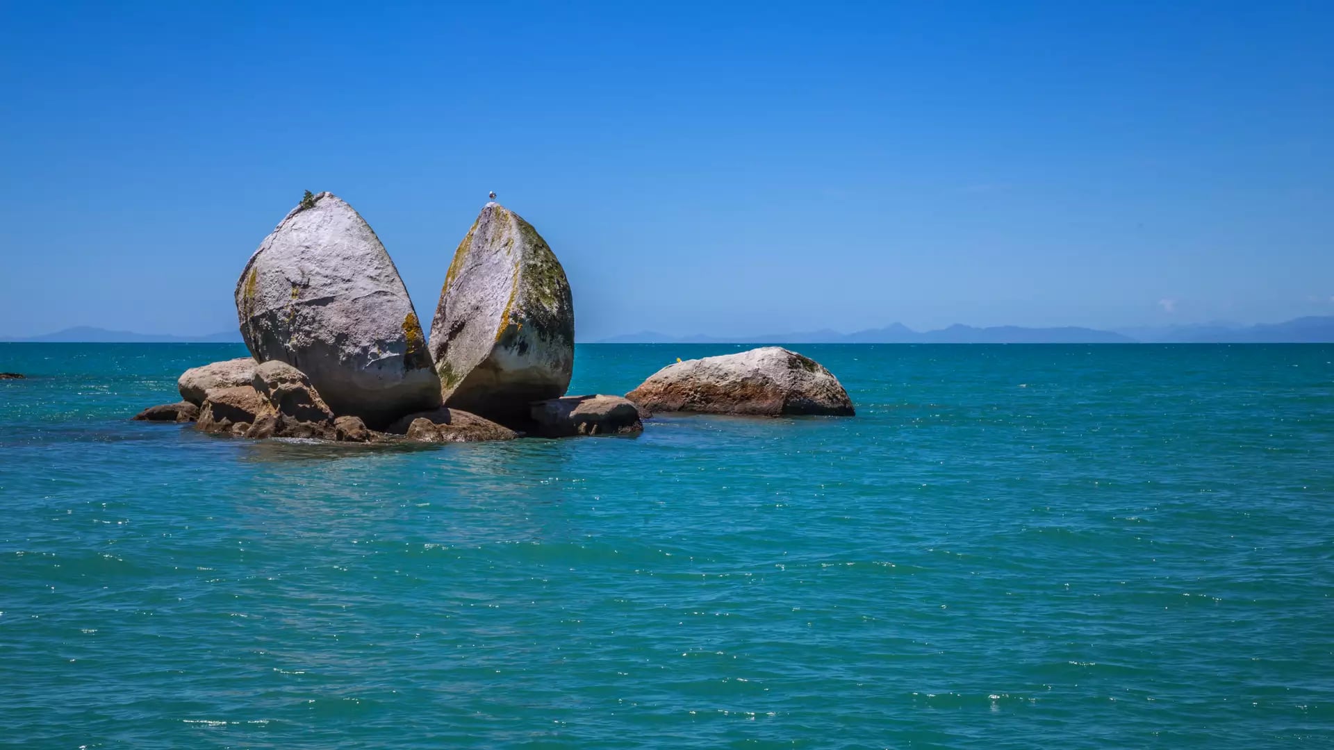 A large split granite boulder in the ocean under a clear blue sky.