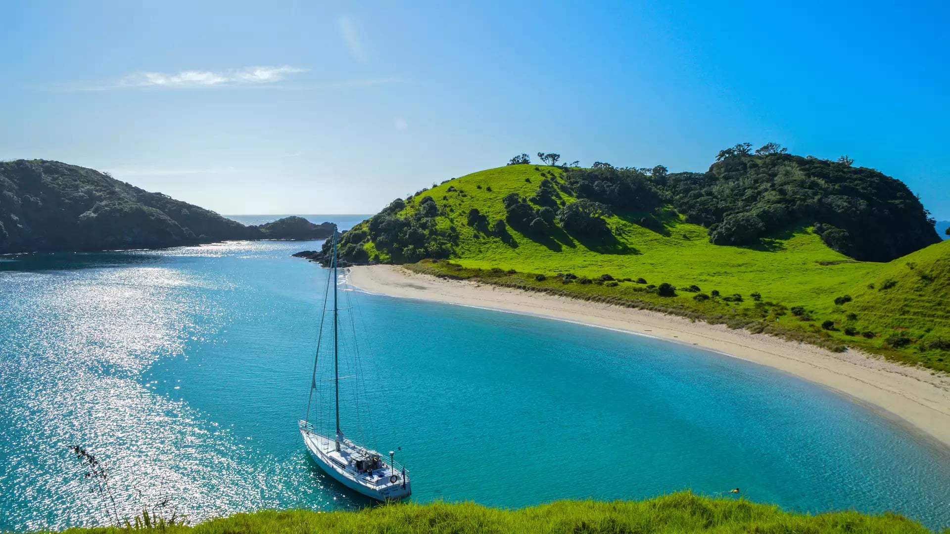 Bright blue sky with green verges, a curved sandy beach and the blue sea with a small boat in the centre 