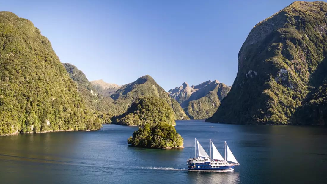 Sailboat navigating a picturesque fjord with green mountains and clear blue sky.