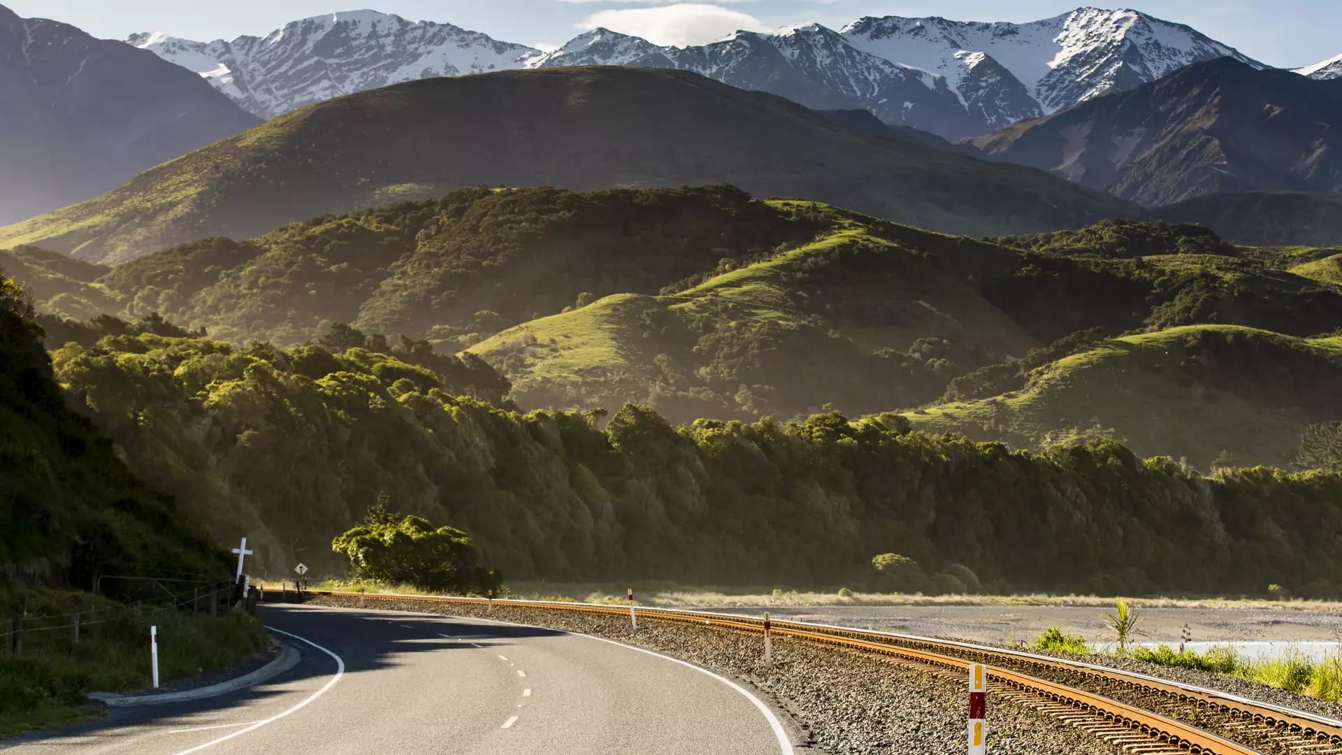 Road and railway track next to green hills with snowy mountains in the background.