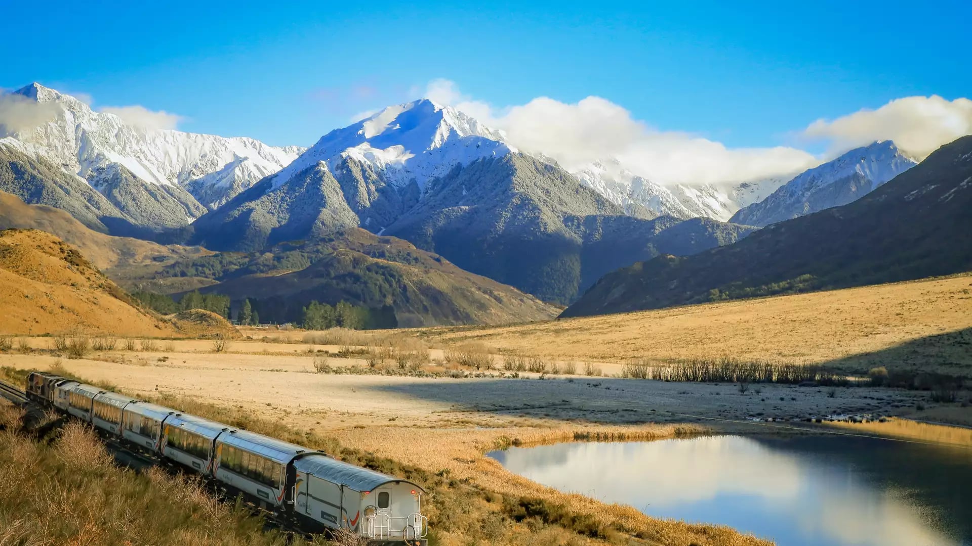 Train passing through valley with snow-covered mountains and lake reflecting sky and clouds