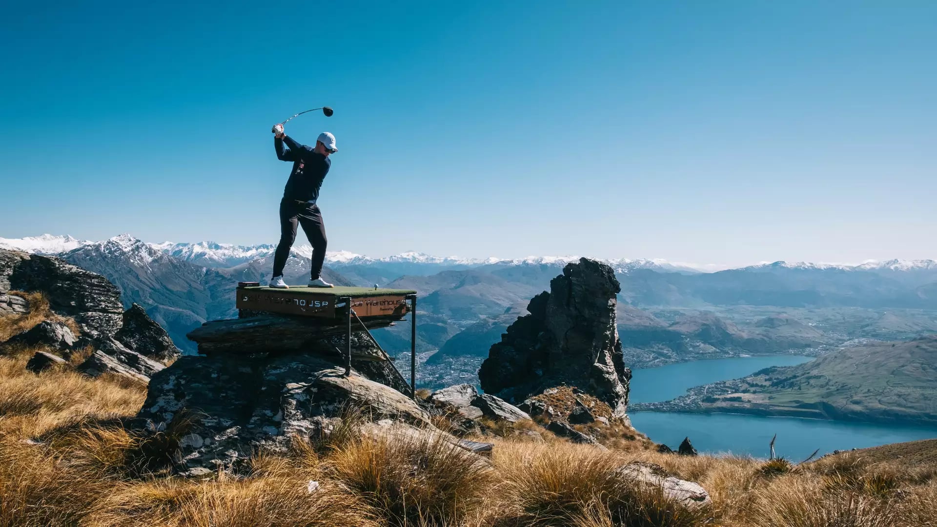 Golfer swinging club on elevated platform with mountains and lake in the background.