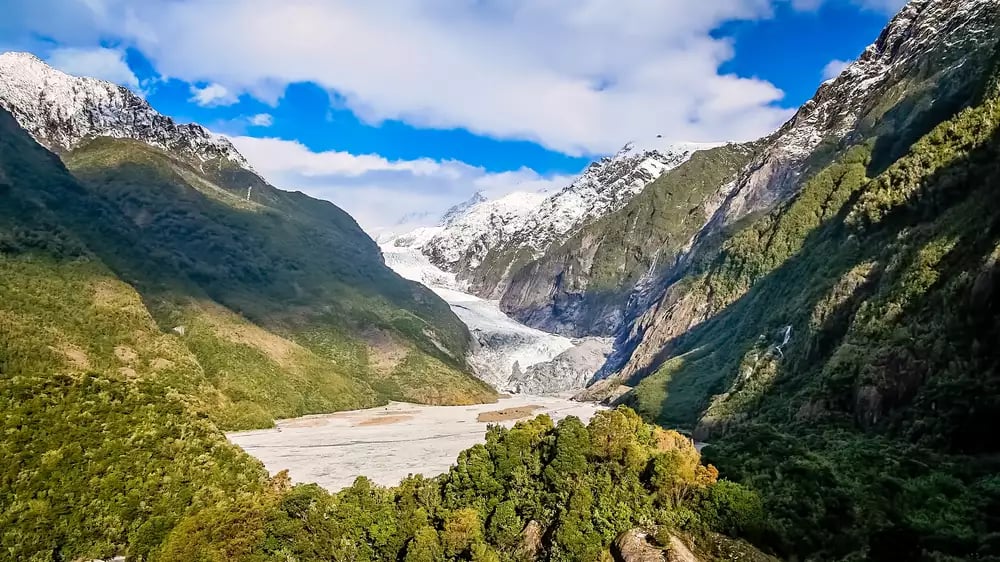 A wide valley view showing the white ice of Franz Josef Glacier descending between steep, green forest-covered mountains under a blue sky with white clouds.