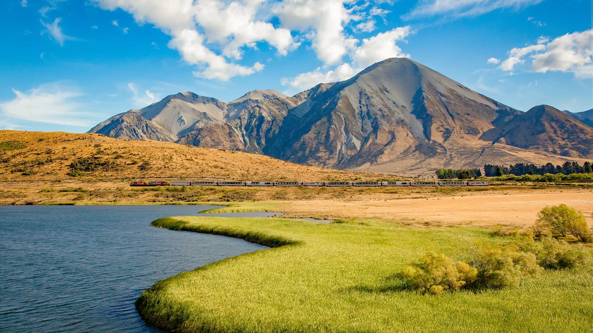 TranzAlpine train passing near Lake Sarah with mountains and blue summer sky