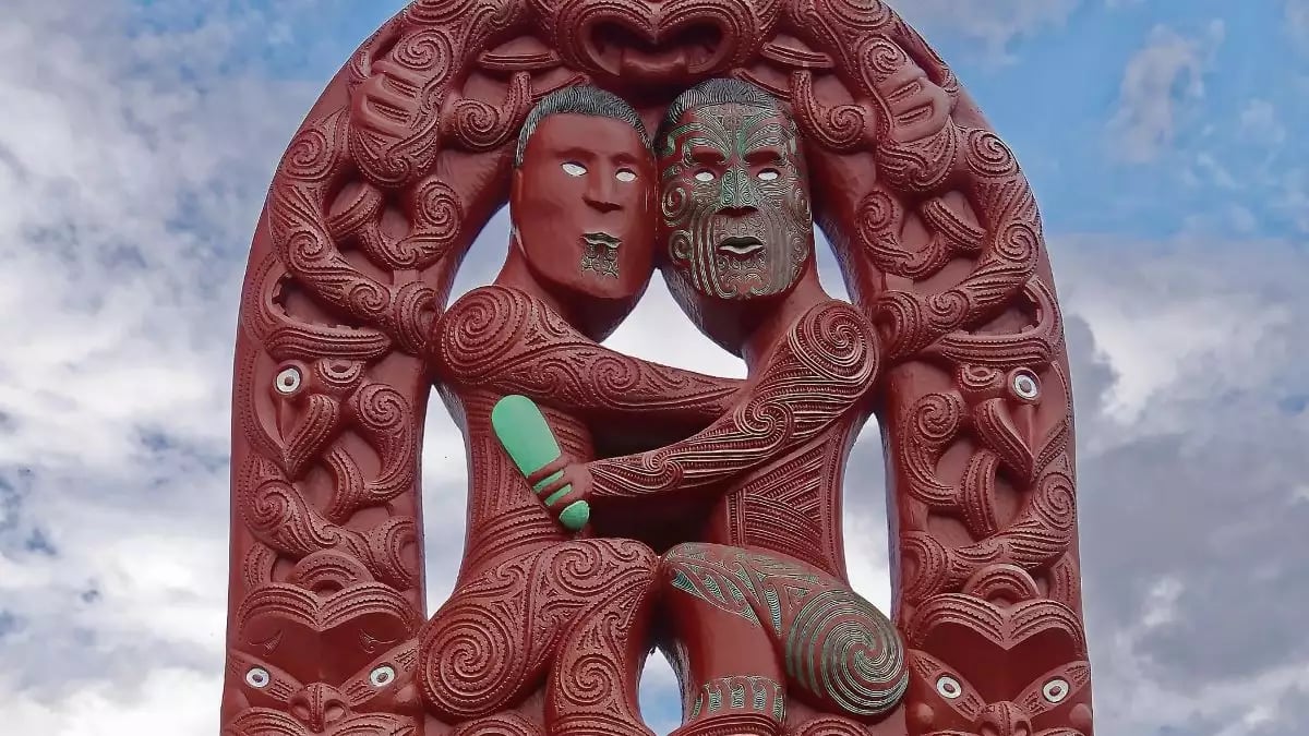Red Maori wood carving of two figures intertwined with intricate patterns under a cloudy sky