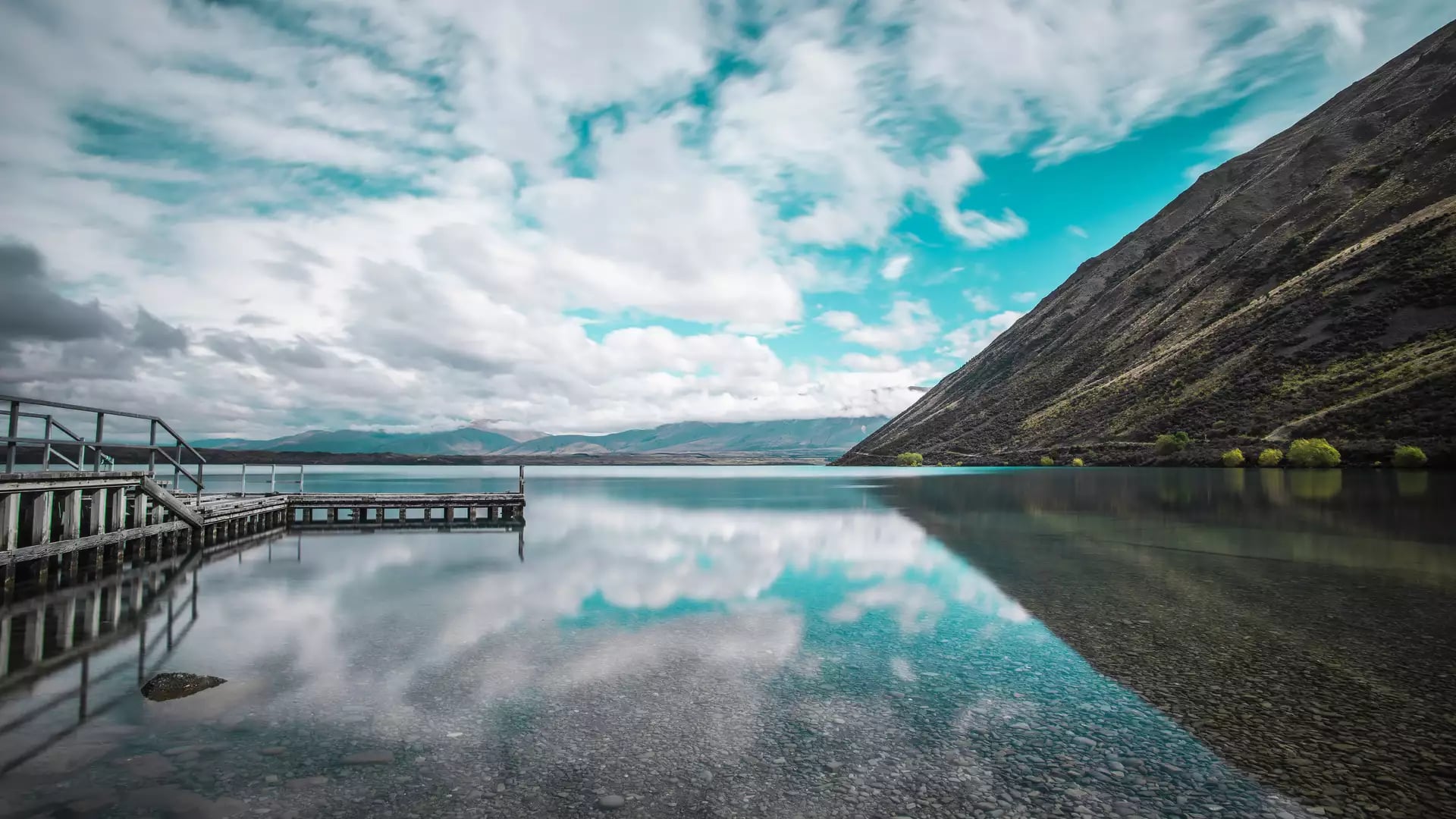 Clear lake with mountain slopes, wooden pier, and clouds reflected in the water at Lake Ohau.