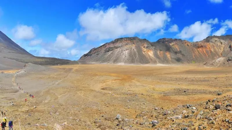 A wide panoramic view of the Tongariro Alpine Crossing showing hikers walking through a barren, rocky volcanic crater with the towering cone of Mount Ngauruhoe in the background under a blue sky.