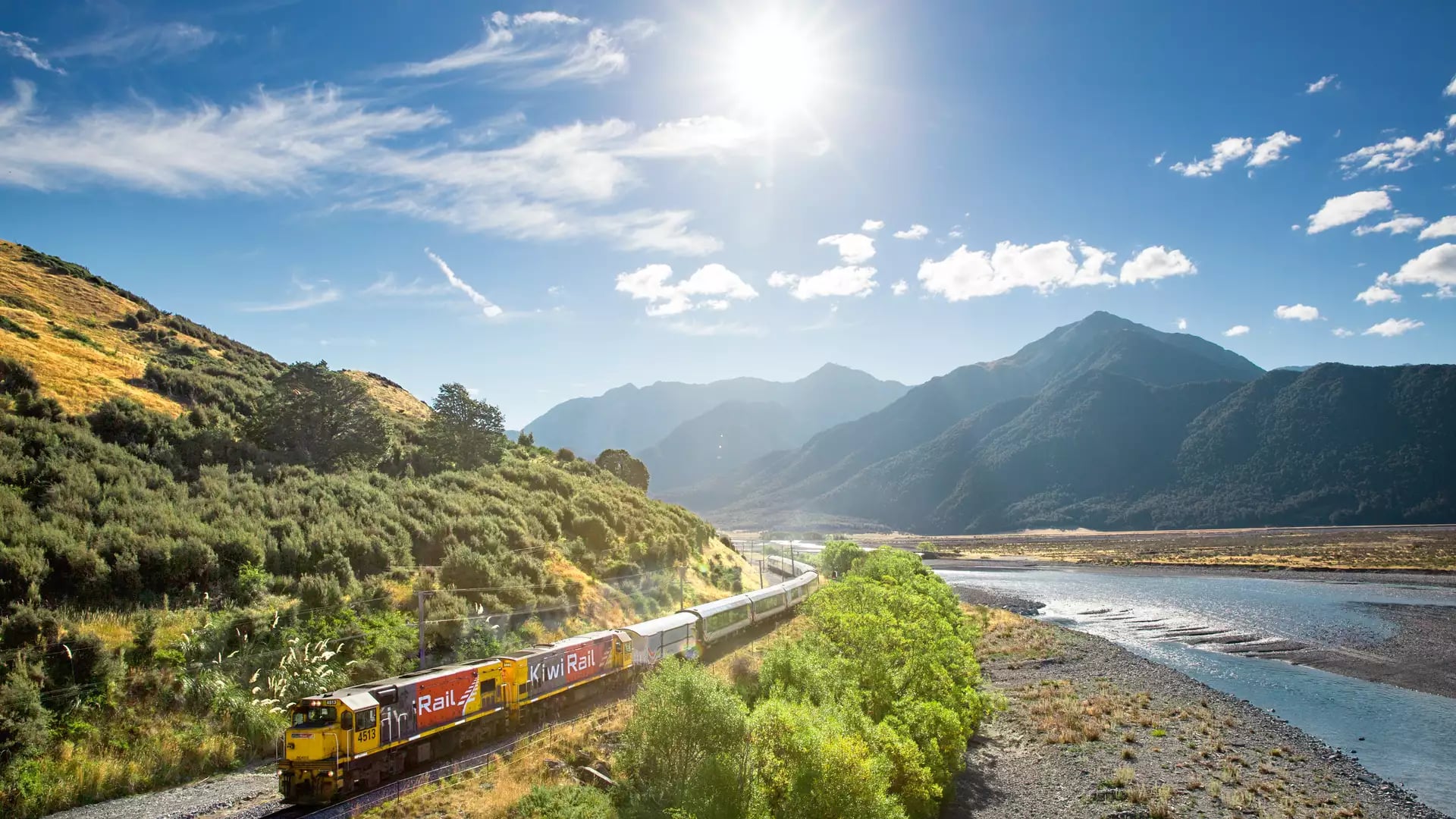 Scenic train crossing a bridge over the Waimakariri River with mountains in the background on a clear day.