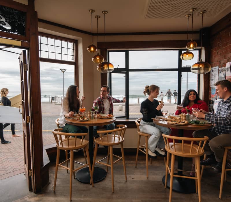People dining inside a seaside café with ocean view and surfers walking outside the door.