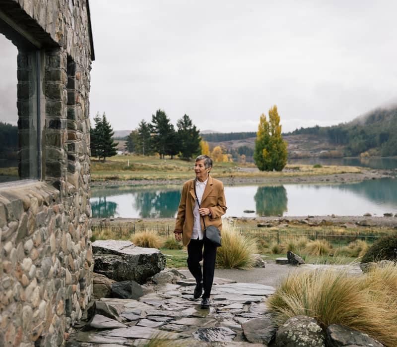 A woman in a tan coat walking along a stone path next to a historic stone church with a misty lake and mountains in the background.