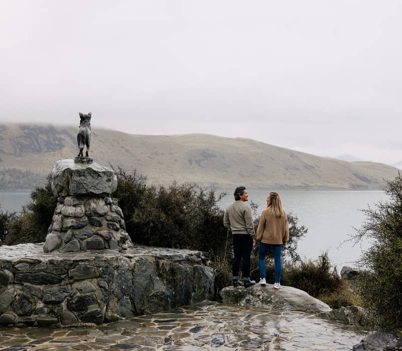 A couple standing on rocks near a bronze statue of a dog on a stone pedestal overlooking a wide, hazy lake and rolling hills.