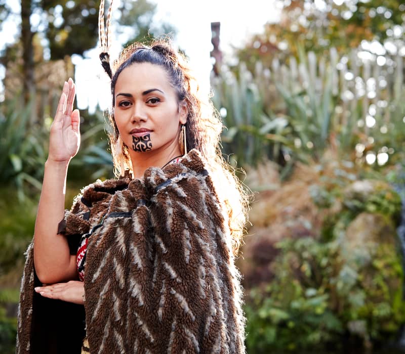 Maori woman with facial tattoo and traditional cloak standing near a waterfall in a forest.
