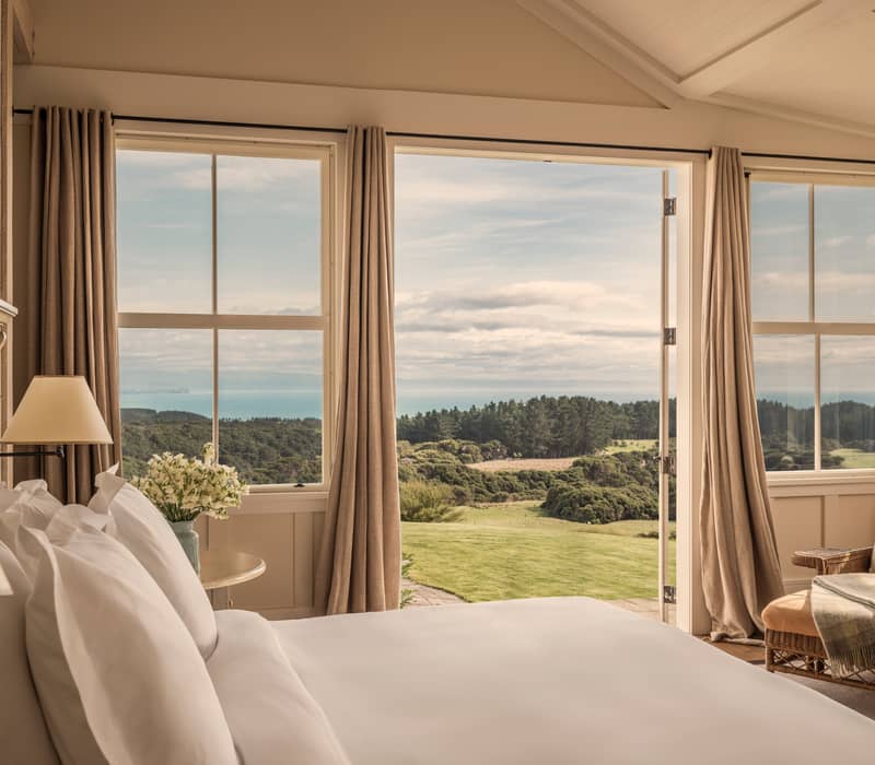 Master bedroom with bed, chair, table, and open door showing scenic green landscape and distant hills.