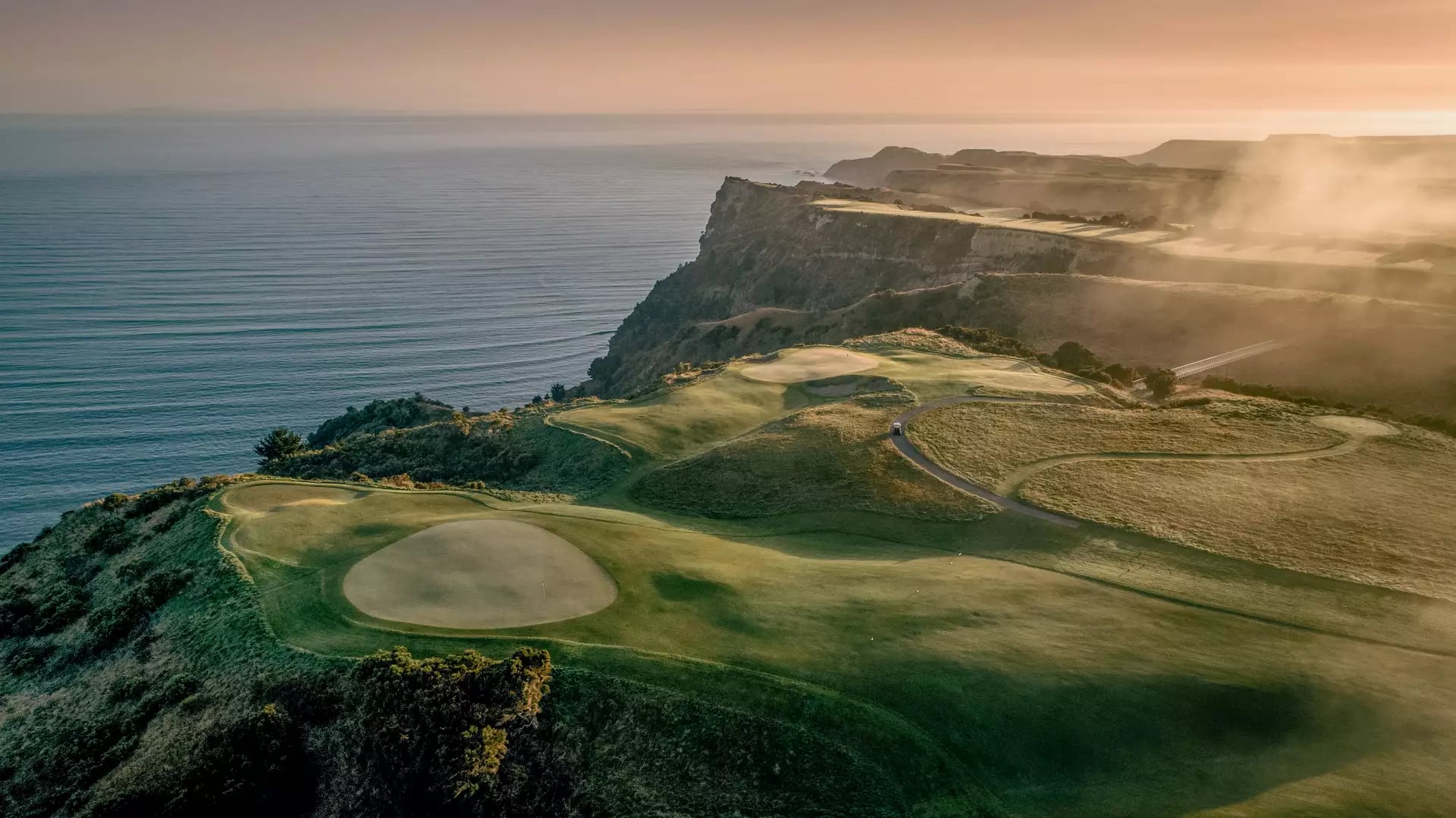 Aerial golf course on cliff by ocean at sunset with green fairways and sand bunkers.