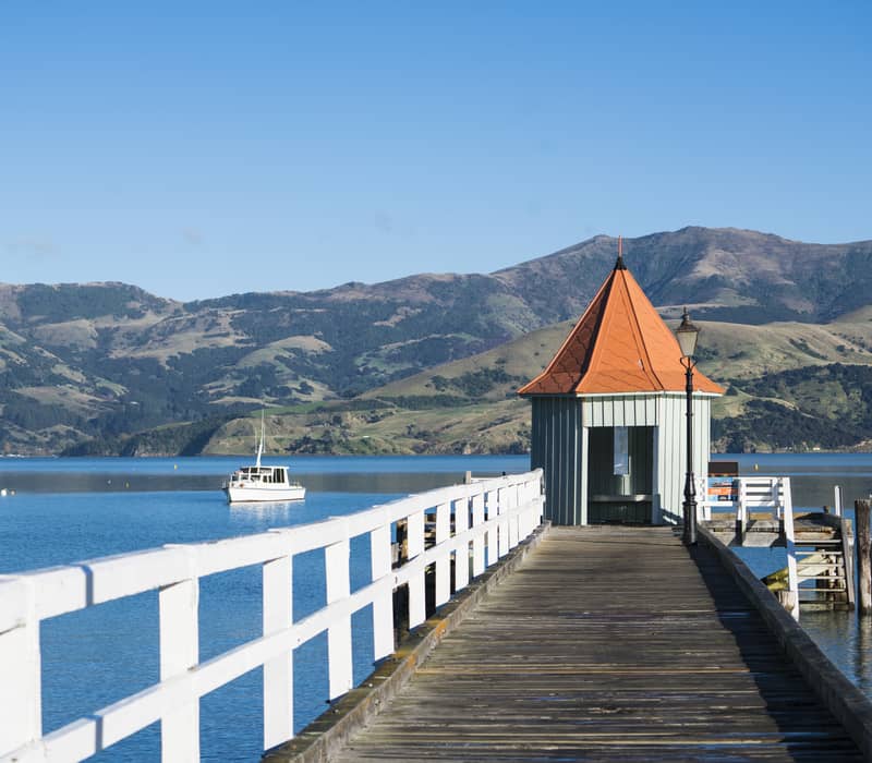 Wooden jetty with a red-roofed shelter extending over calm water with mountains in the background.
