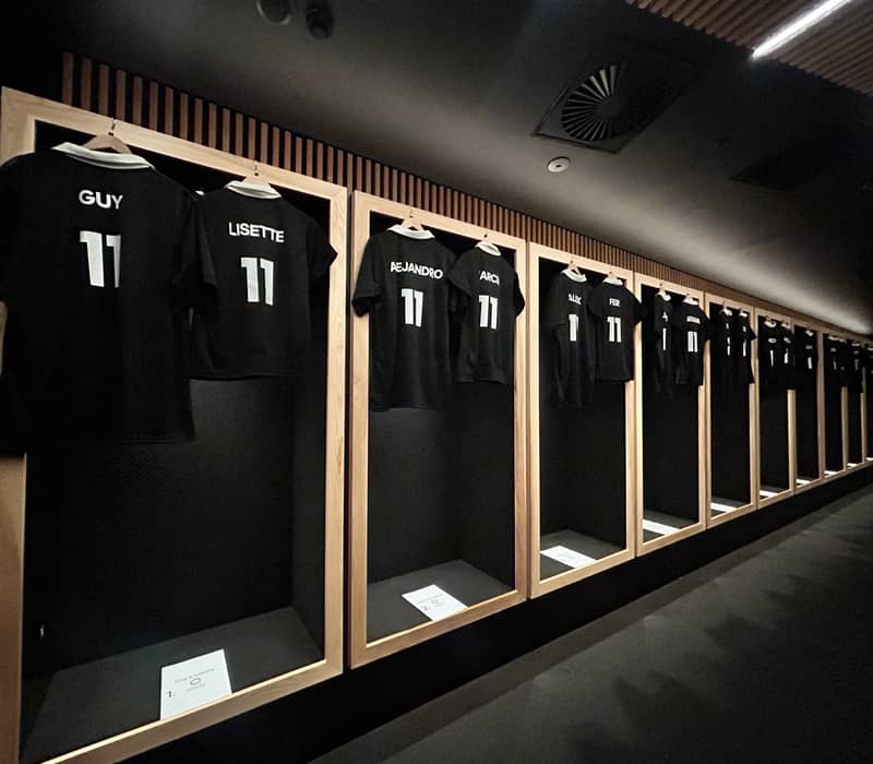 Black sports jerseys with names hanging in wooden framed lockers in a dimly lit changing room.