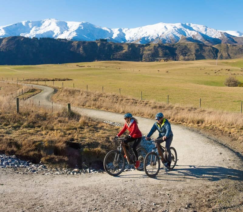 Couple biking on a rural trail with snowy mountains in the background under a clear sky.